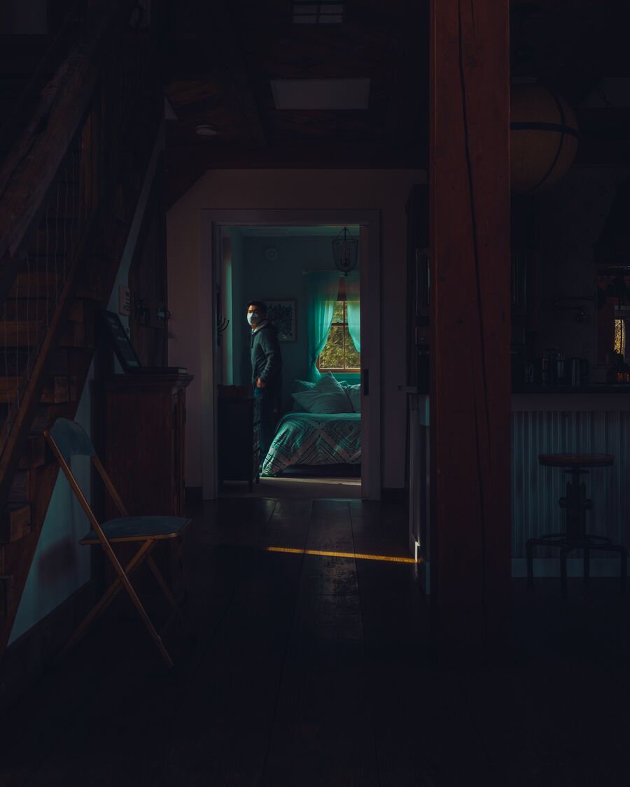 A dark barn interior with a staircase on the left and a timber post on the right. Through a far doorway, a man in a face mask stands in a bedroom lit blue-green.