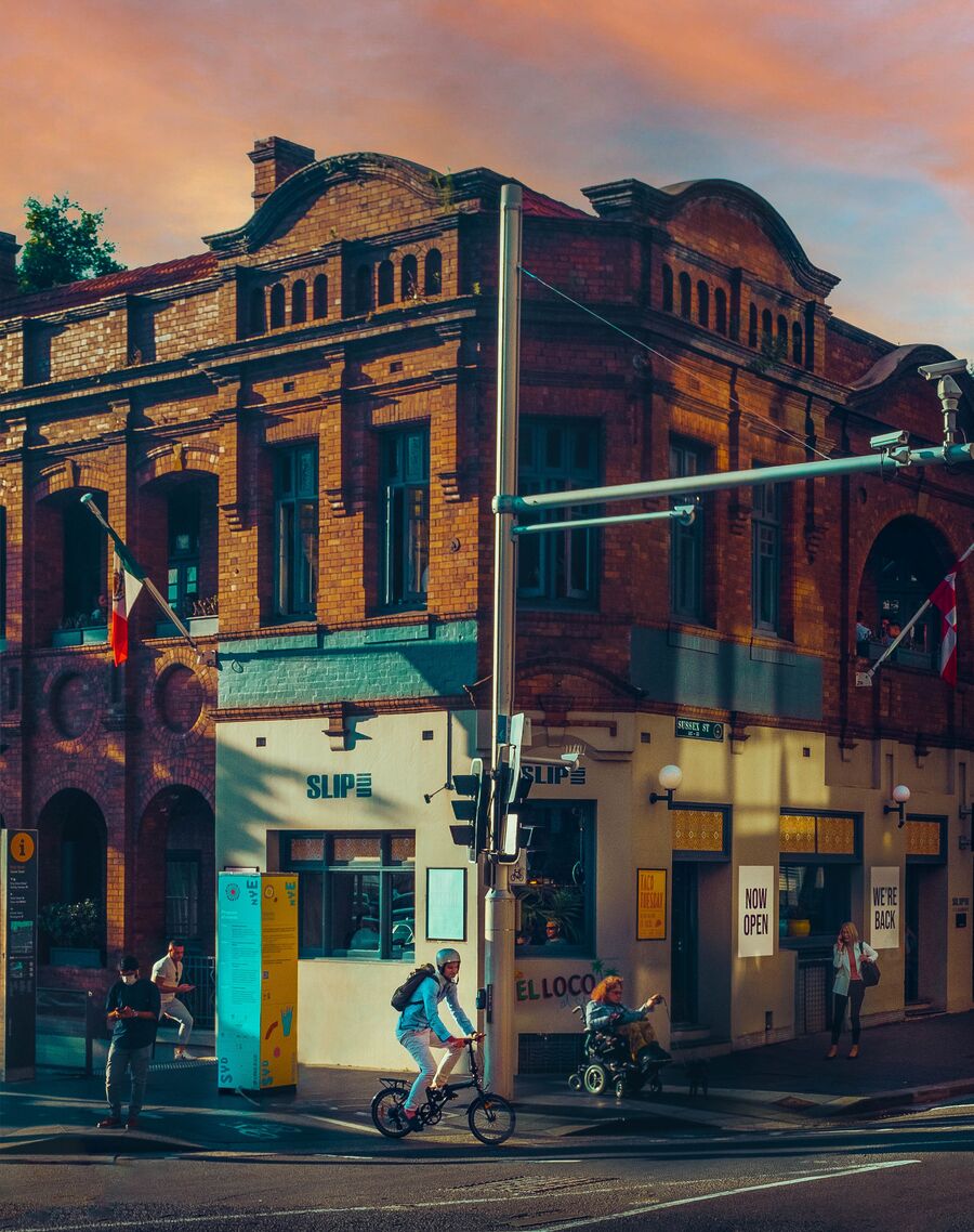 A two-storey brick pub on a Sydney corner in late sun, as a cyclist, a person in a wheelchair and people on foot pass in front.