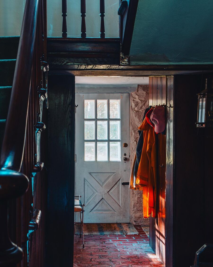 Coats and caps hang on hooks beside a white cottage door with nine glass panes, and low sun lights up an orange raincoat.