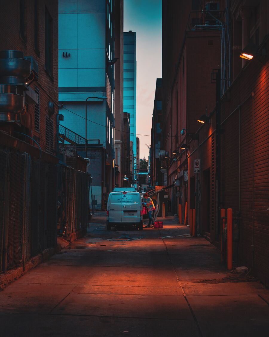 A white grocery van stands in a narrow downtown alley at first light. A worker unloads beside its open side door, and lamps glow on the brick walls.