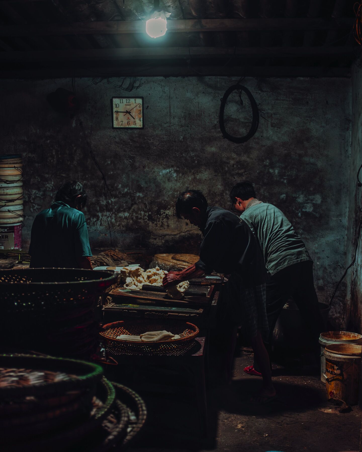 Three men work dough at a long table under a single bulb in a noodle workshop in Hội An. A clock hangs on the soot-dark wall above them.