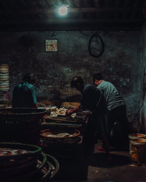 Three men work dough at a long table under a single bulb. A clock hangs on the soot-dark wall above them.