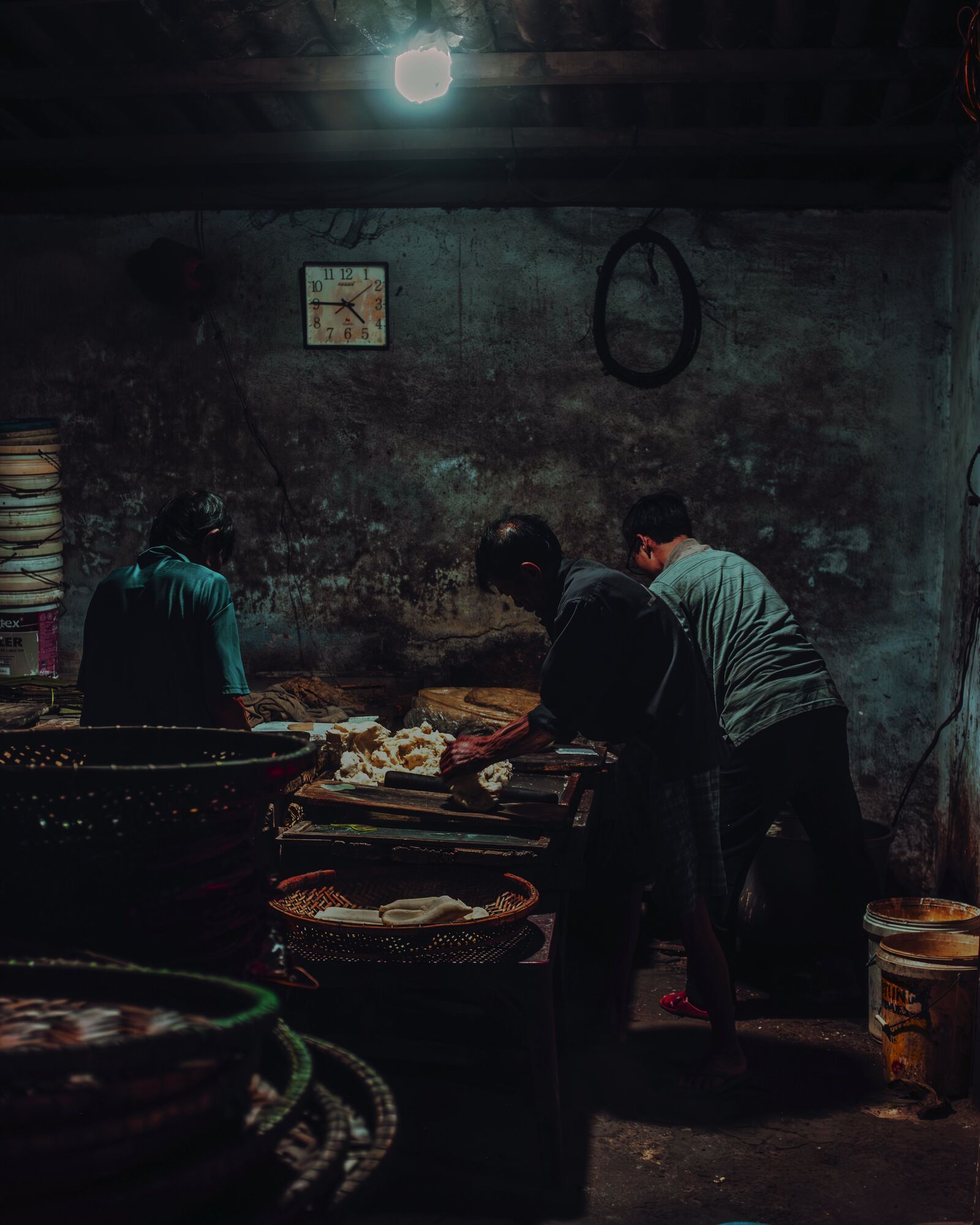 Three men work dough at a long table under a single bulb. A clock hangs on the soot-dark wall above them.
