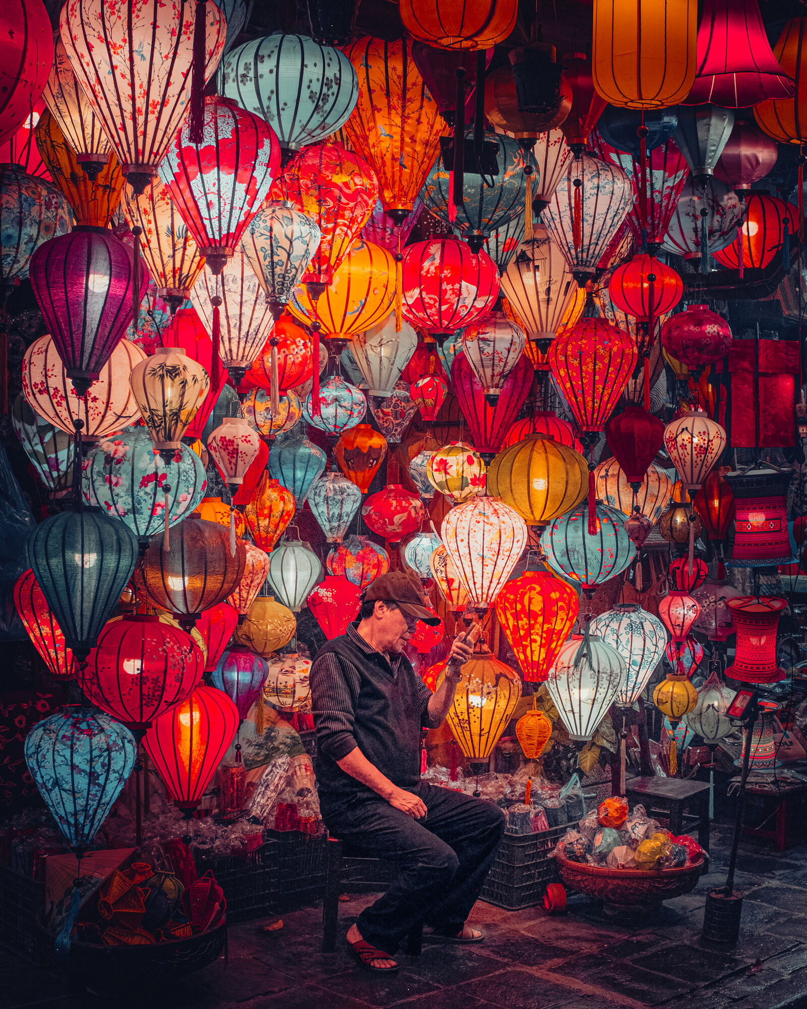 A man in a cap sits on a stool in front of a wall of lit silk lanterns, looking down at the phone in his hand. A basket of wrapped lanterns sits on the wet paving beside him.