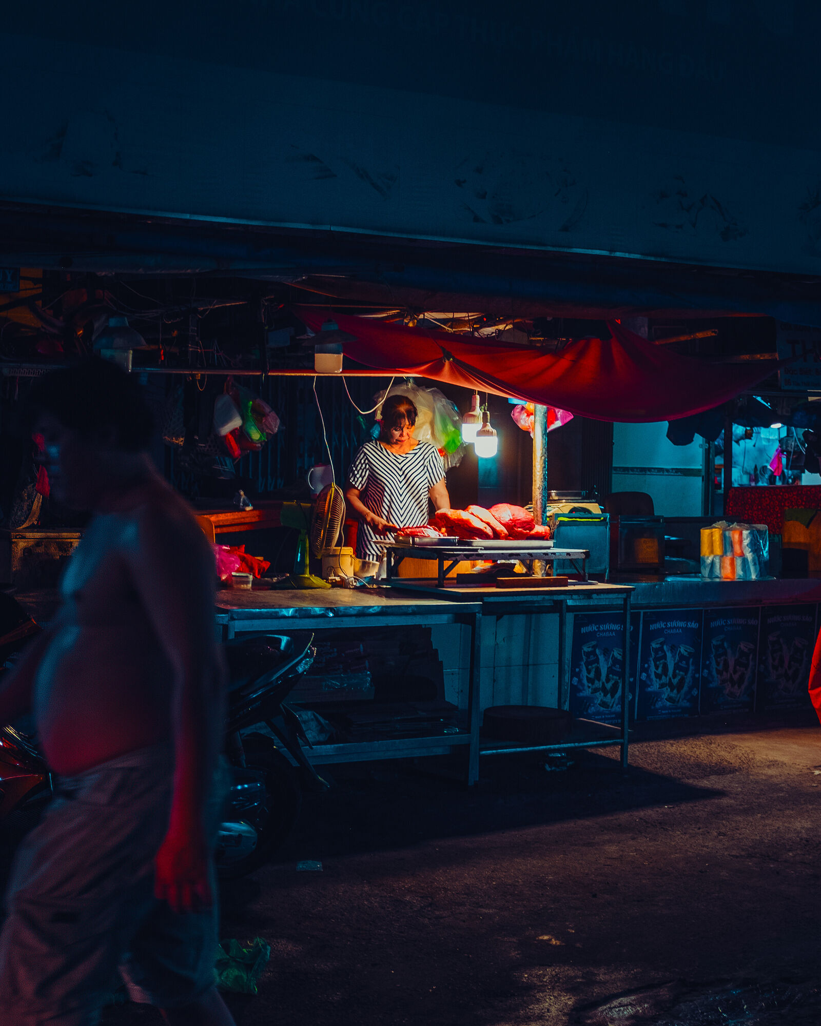 A woman in a striped dress works over large cuts of beef at a market stall, under hanging bulbs and a red tarp. A shirtless man, blurred and washed in red light, walks through the left of the frame.