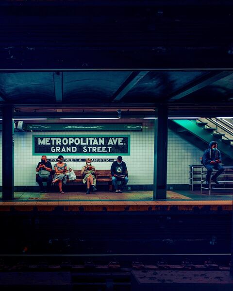 Seen across the tracks, four masked people sit on a wooden bench under a tiled sign reading Metropolitan Ave. Grand Street, two on their phones and two reading. A fifth sits on a railing by the stairs.