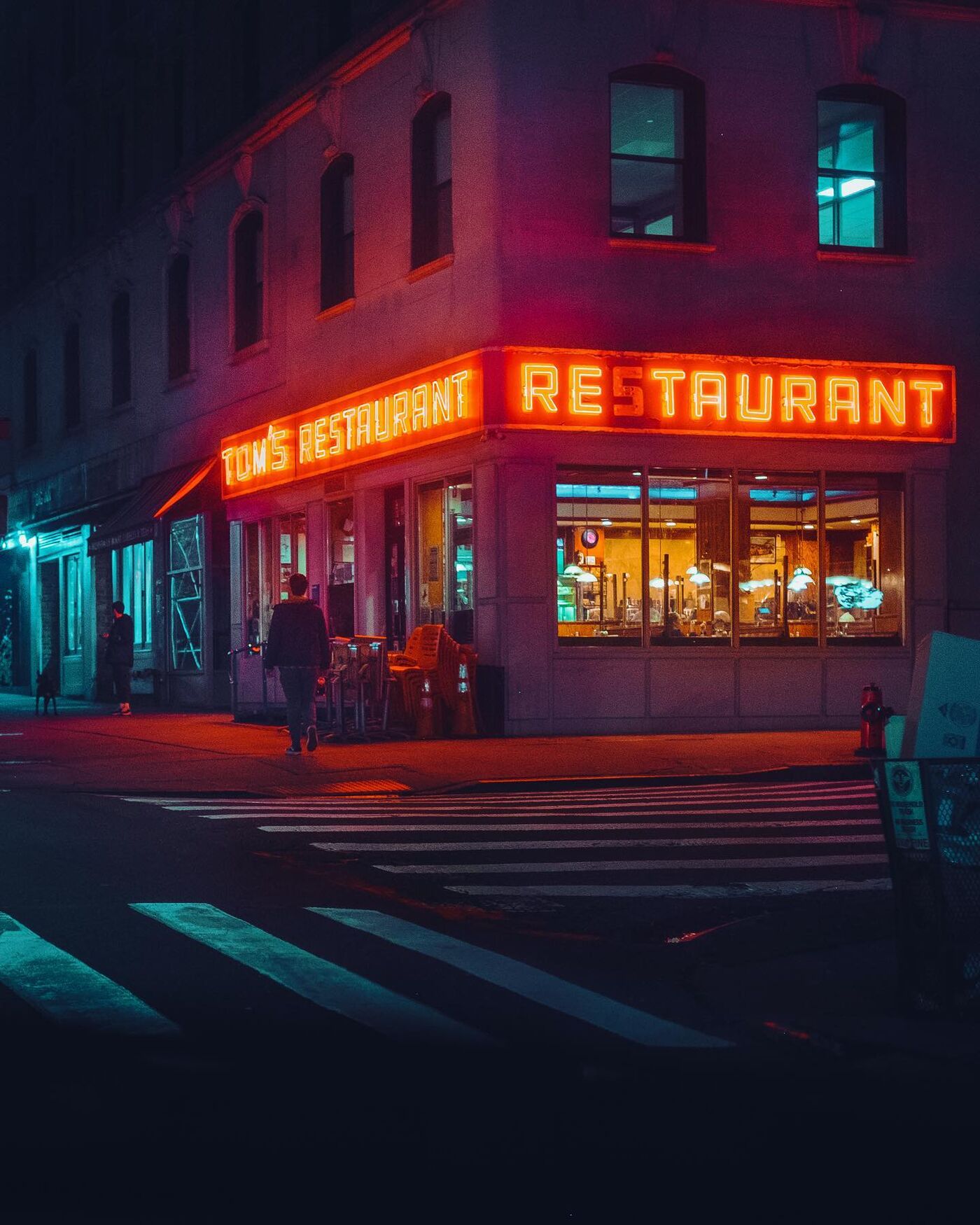 Tom's Restaurant on its corner at night, its red neon reading Tom's Restaurant on one side and Restaurant on the other. A man walks past stacked chairs out front, the crosswalk is empty, and further down the block someone stands with a dog.