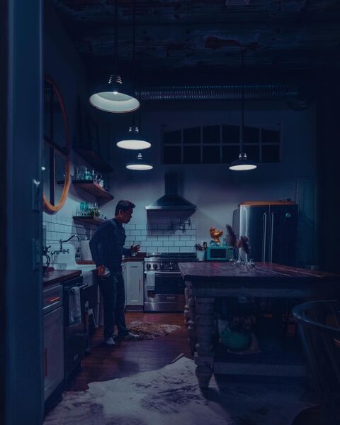 A man stands with a glass of water in a dark kitchen under four pendant lamps, beside a farmhouse sink and white subway tile. In the foreground, a long wooden table on turned legs stands over a cowhide rug.