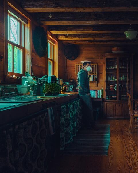 A man in a grey hoodie and a white face mask leans on a long wooden counter in a timber cabin kitchen, hands in his pockets. Daylight comes through tall windows, with bare branches outside.