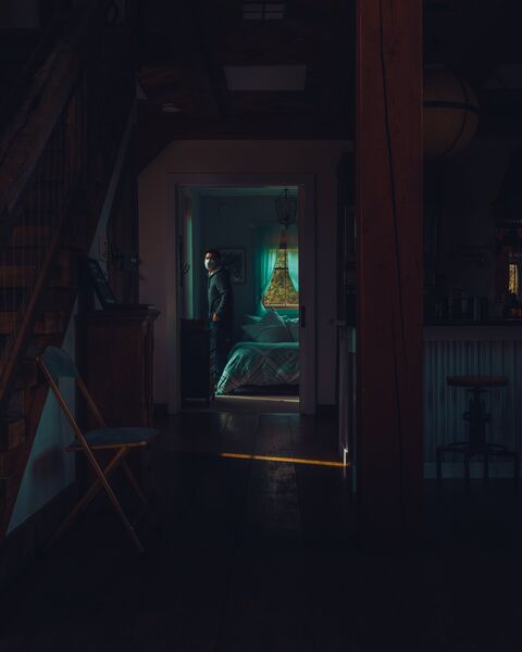 A dark barn interior with a staircase and a folding chair on the left and a timber post on the right. Through a far doorway, a man in a face mask stands in a bedroom lit blue-green, with trees in the window behind him.