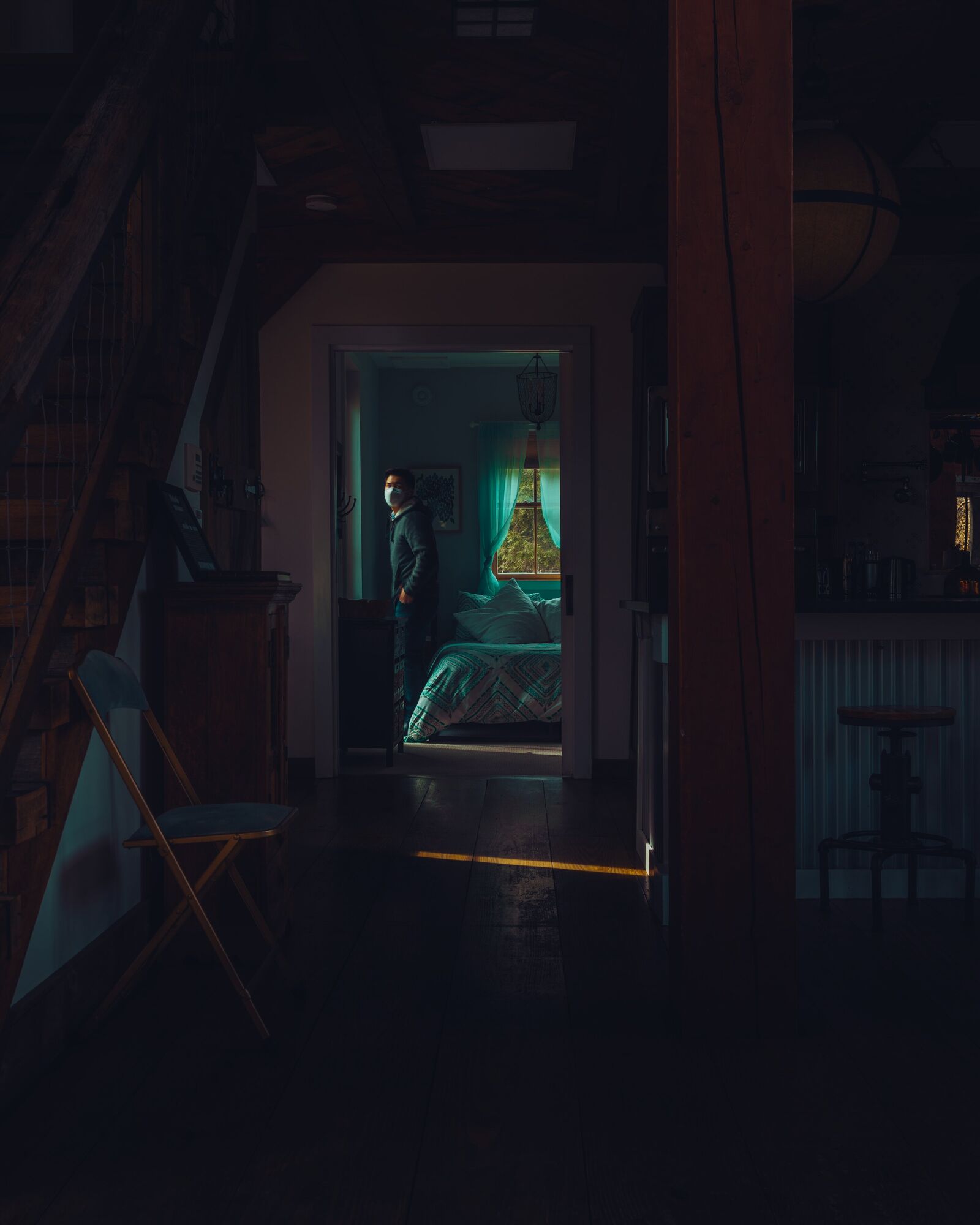 A dark barn interior with a staircase and a folding chair on the left and a timber post on the right. Through a far doorway, a man in a face mask stands in a bedroom lit blue-green, with trees in the window behind him.