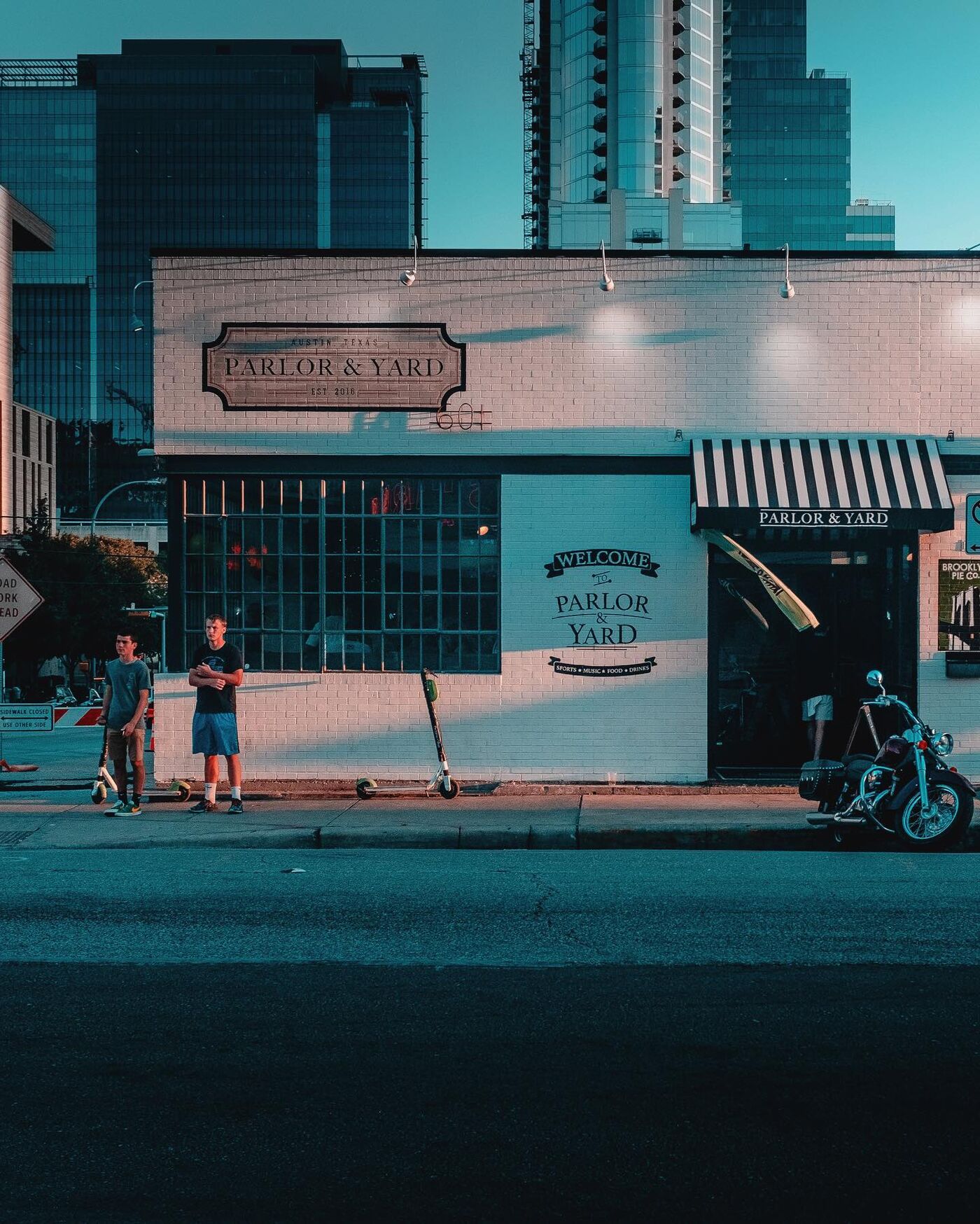Two teenagers with scooters stand at the kerb by Parlor & Yard's white brick wall. A motorcycle is parked by the door under a striped awning, and glass towers rise behind.