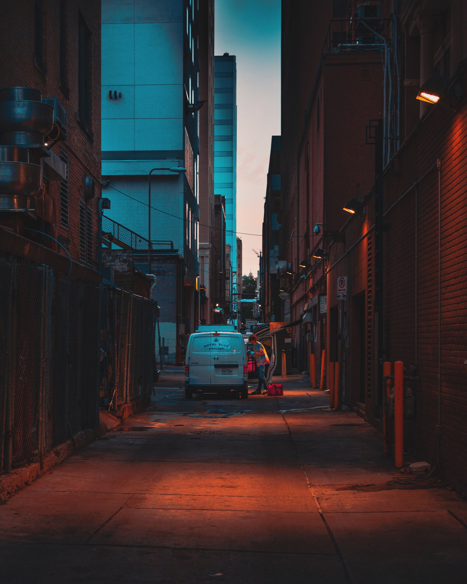 A white Royal Blue Grocery van stands in a narrow downtown alley at first light. A worker unloads beside its open side door, and lamps glow on the brick walls.