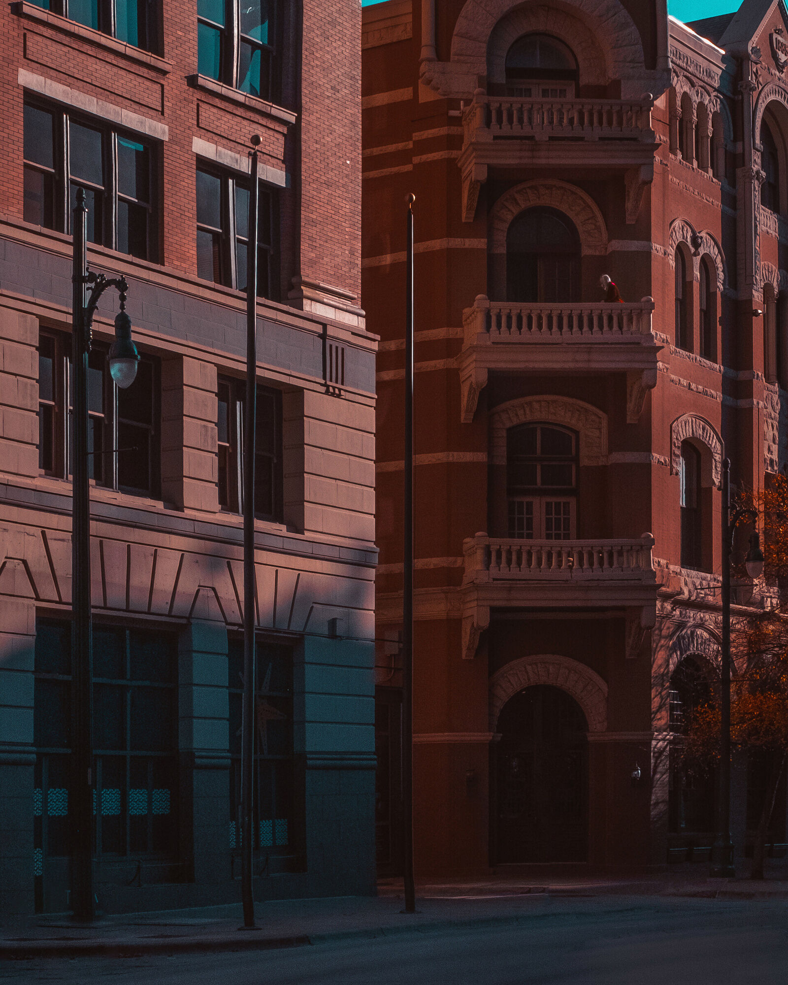 Low sun on The Driskill's red-brick front, where three stone balconies stack above an arched doorway. A person stands on the middle balcony. Lamp posts line the empty street beside a second brick building.